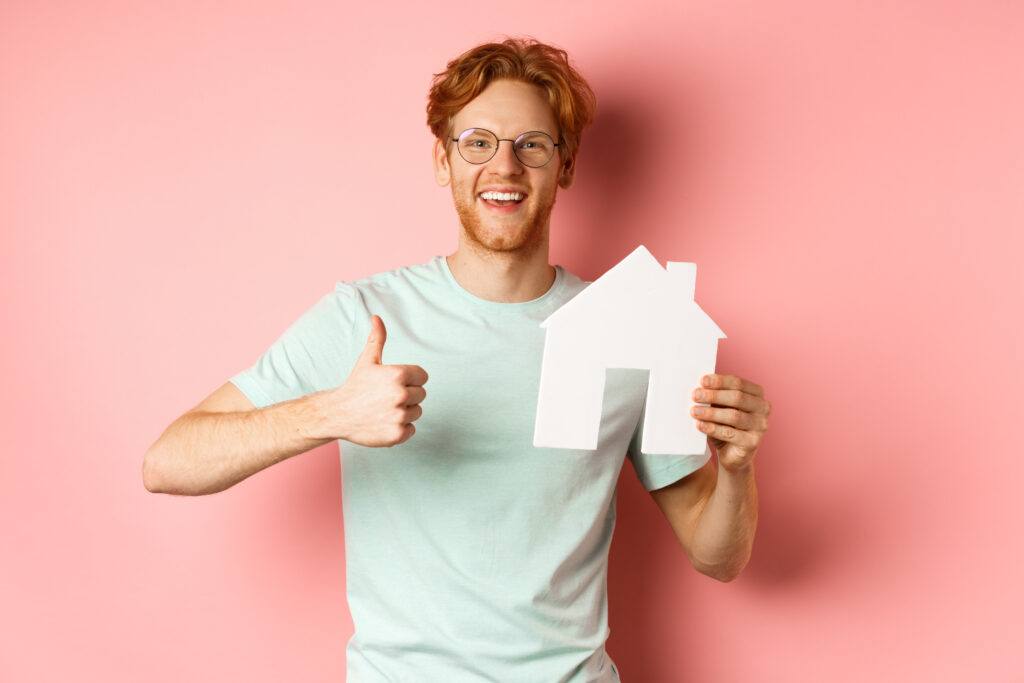 Real estate. Cheerful man in glasses and t-shirt recommending broker agency, showing paper house cutout and thumbs-up, standing over pink background.